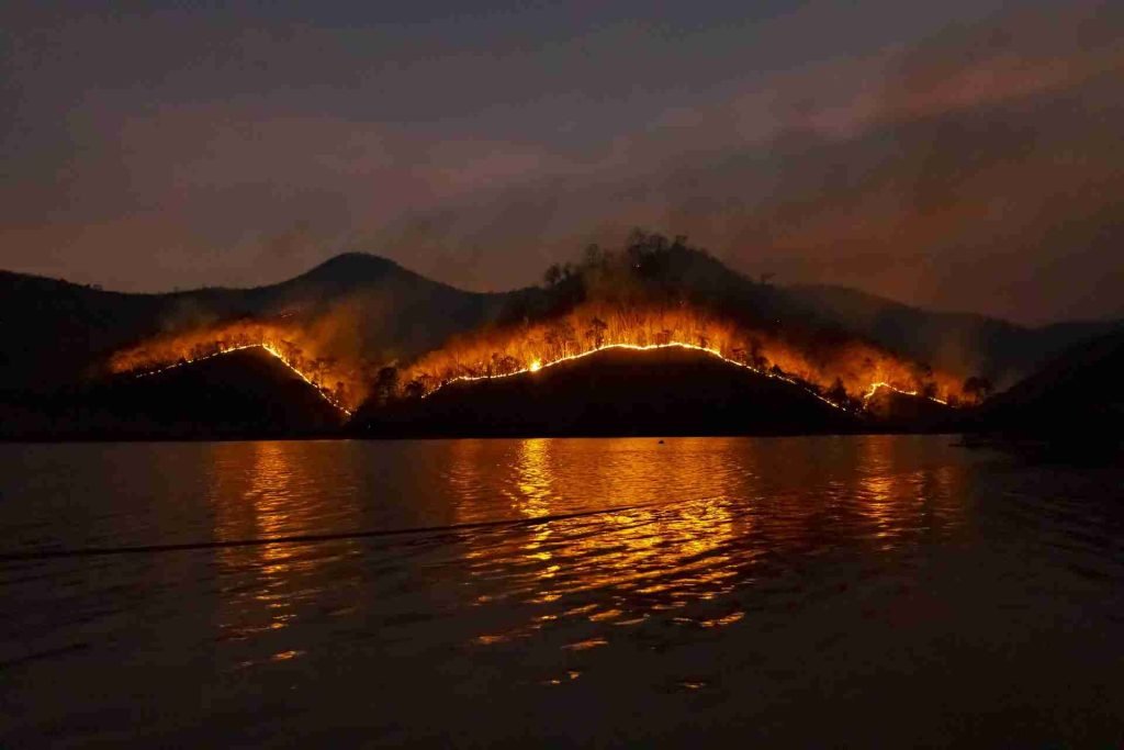 Wildfire burning on hills reflected in a calm body of water at night.
