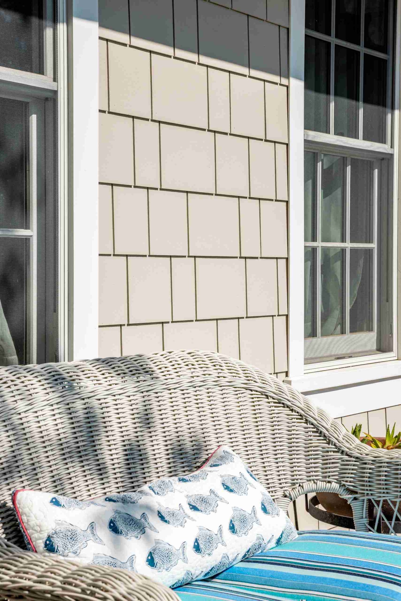 All Hail Fiber Cement: How Fiber Cement Siding Protects Homes 4 Wicker bench with a blue striped cushion and a pillow featuring fish designs against a beige shingled wall with windows.