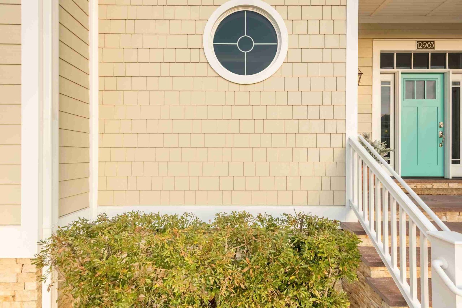 Front of a house with a circular window, teal door, and a white railing leading to the entrance, surrounded by green shrubbery.