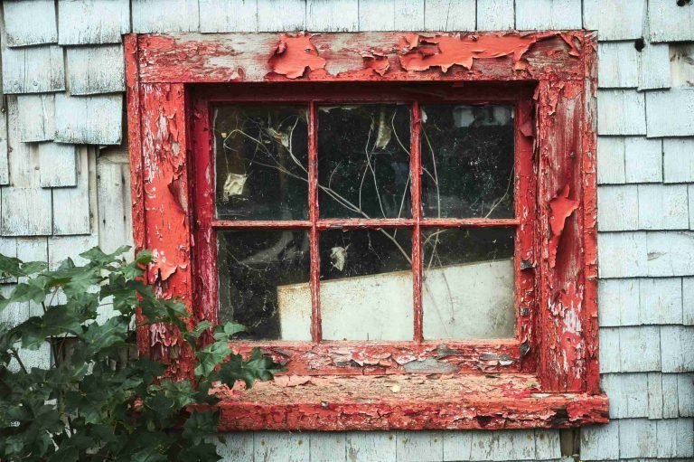 Weathered red window frame with peeling paint and cracked glass set in a white shingled wall, partially obscured by greenery.