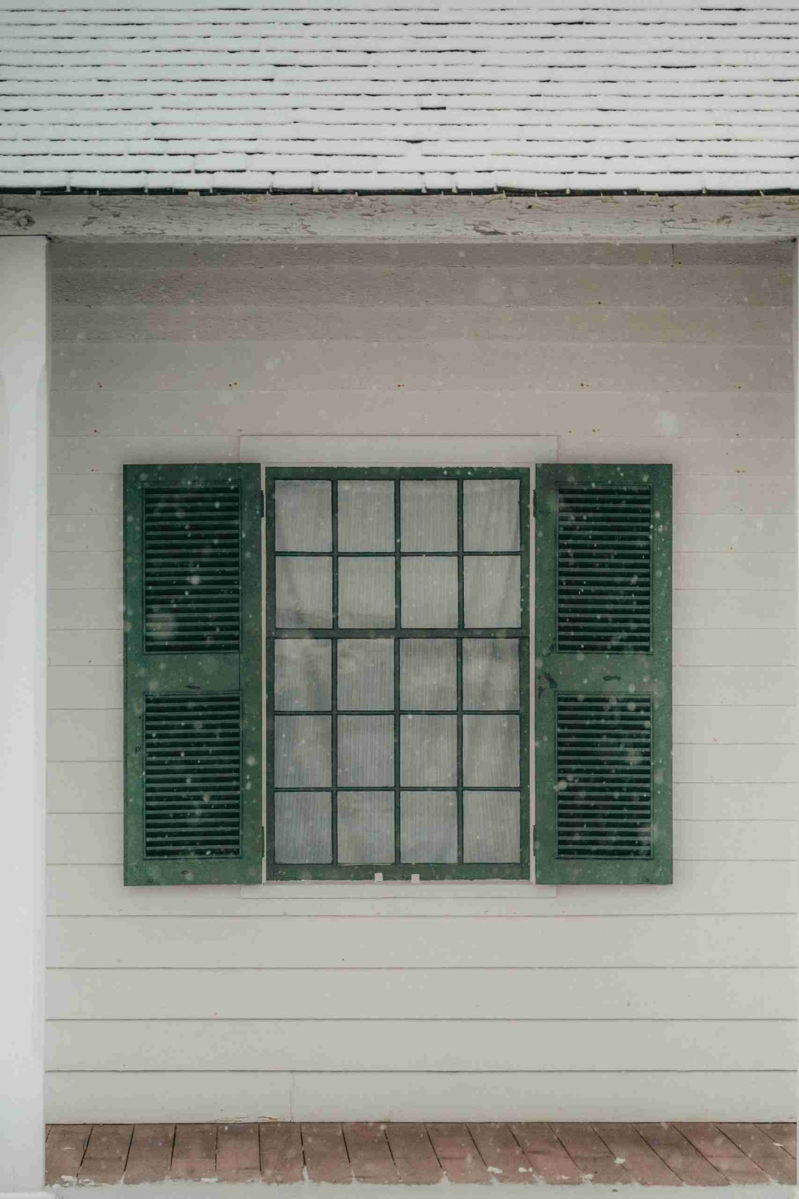 The Pain of Old Panes: How Old Windows Can Cost You 2 A single window with dark green shutters on a gray wooden house wall, with a snowy roof visible above.