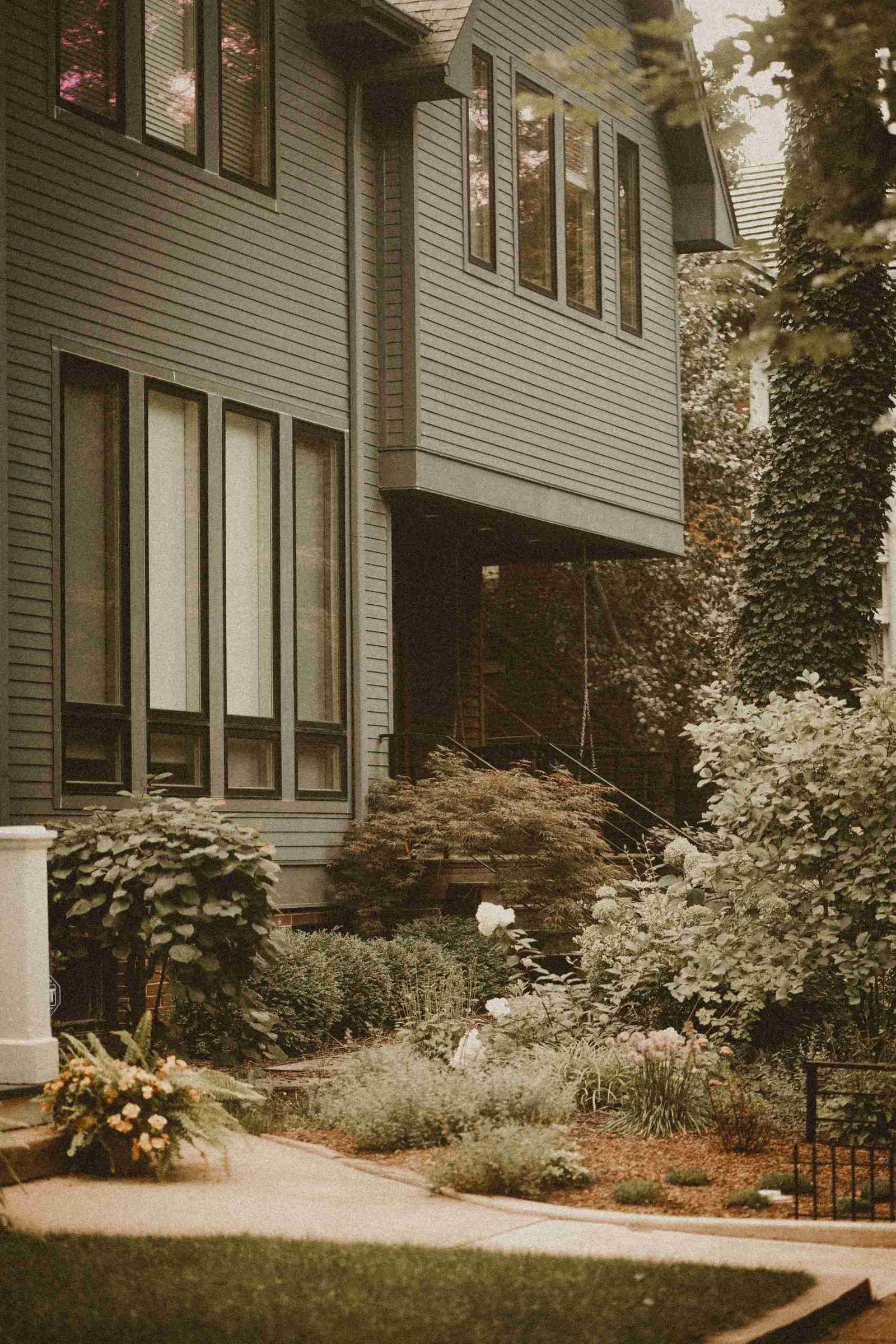 A dark gray house with large windows and a lush, well-maintained garden in the foreground, featuring various plants and shrubs.
