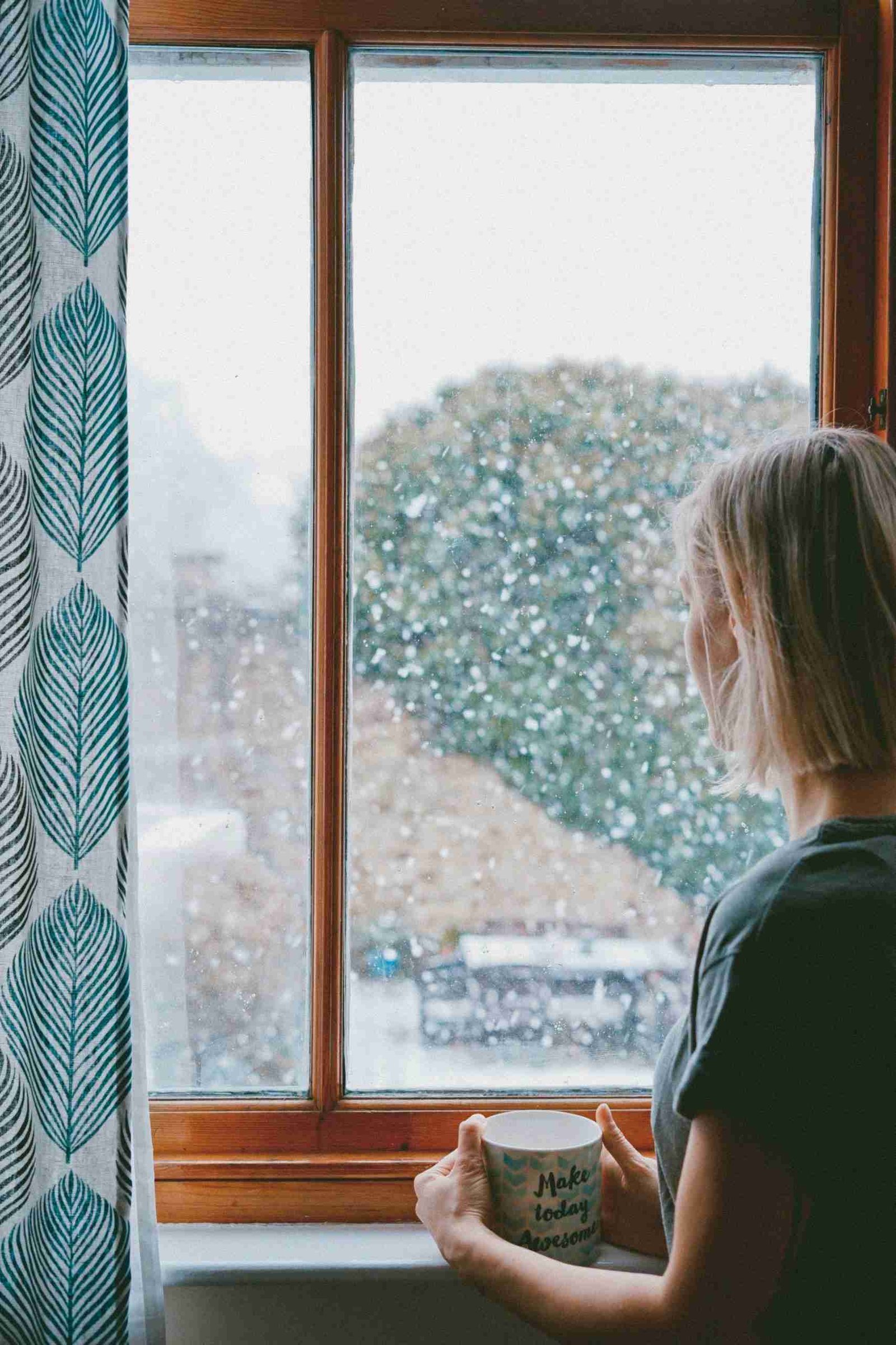 The Pain of Old Panes: How Old Windows Can Cost You 3 A person stands by a window holding a mug, looking outside at a snowy scene with trees in the background.